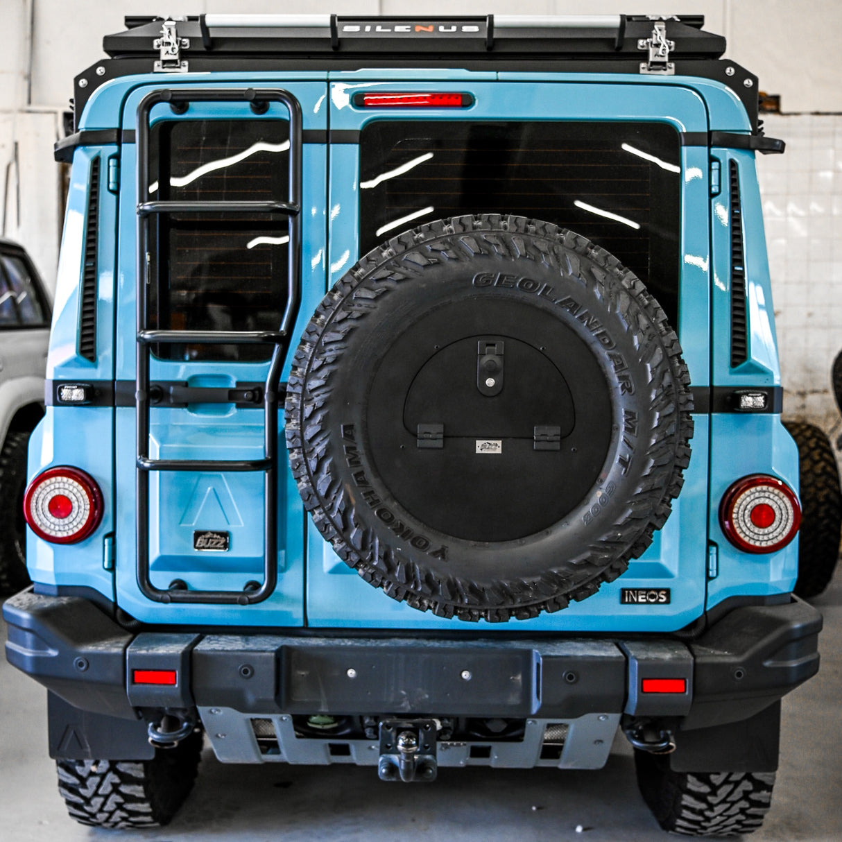 Buzz Overland Spare Wheel Storage Cubby for INEOS Grenadier, made from corrosion-resistant 5052 aluminium with a textured black powder coat, bolted to a 17-inch wheel for secure off-road storage.